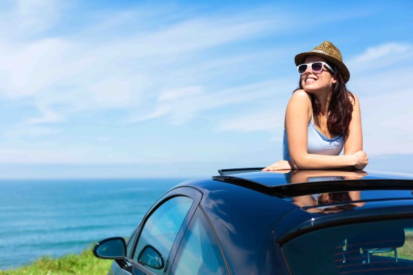 Relaxed happy woman on summer travel vacation to the coast leaning out car sunroof with the sea on background.